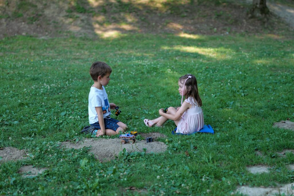 Two young children sitting on the grass, facing each other and playing with toy vehicles in a park.